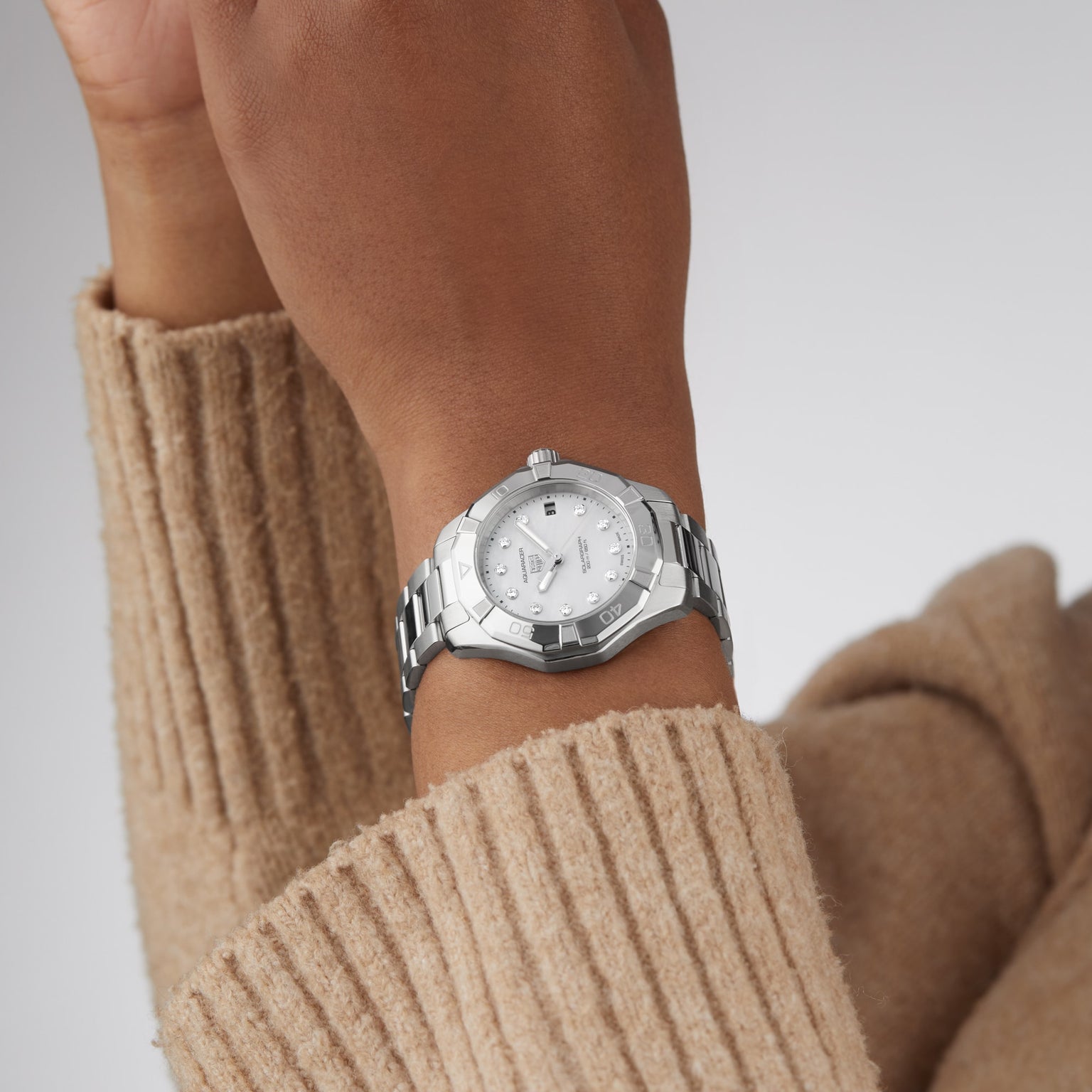 A person wears a TAG Heuer Aquaracer 200 Professional Solargraph silver sports watch with a white dial and stainless steel band, shown against a tan sweater sleeve.