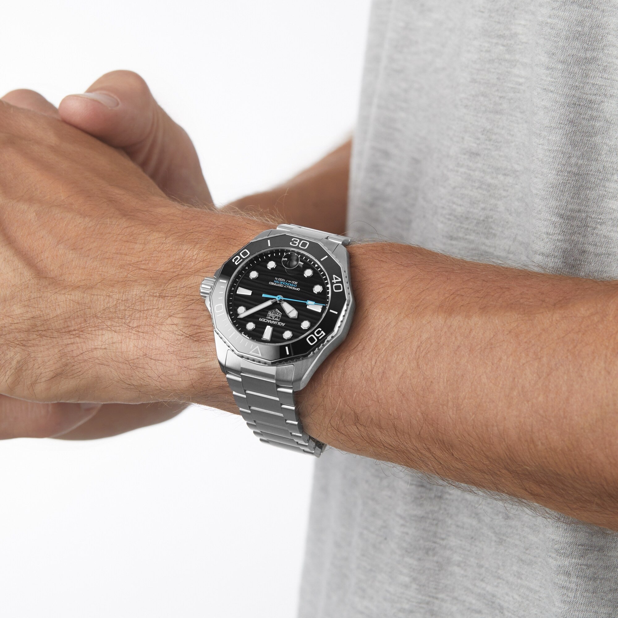A person wears a TAG Heuer Aquaracer Professional 300 Date with a black dial and rotating bezel on their left wrist; the silver stainless steel watch stands out against a light background.
