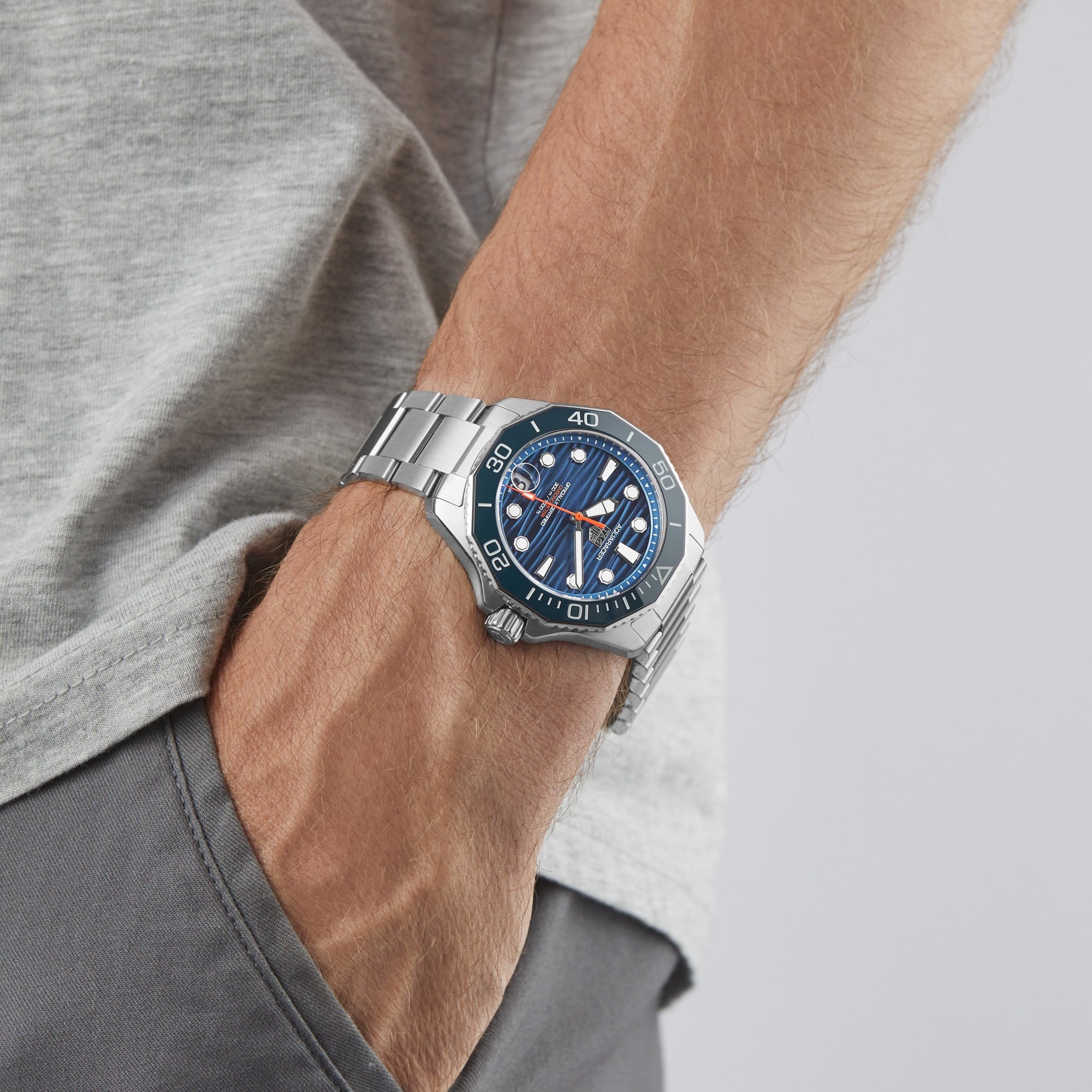 A person wearing a TAG Heuer Aquaracer Professional 300 Date with a blue dial and orange accents stands with a hand in the pocket of gray pants—a striking example of Swiss-made craftsmanship.