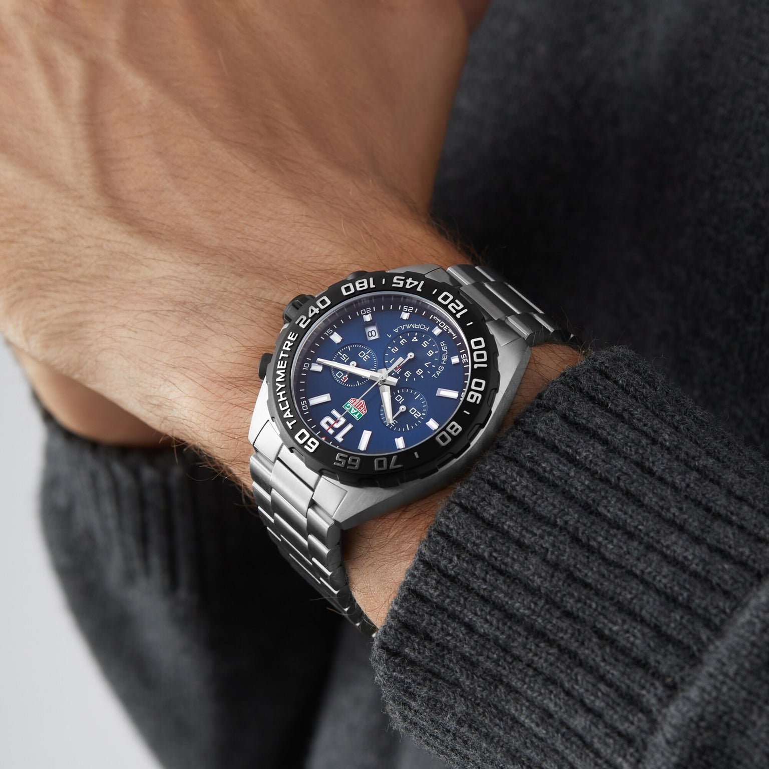 A person wearing a TAG Heuer Formula 1 Chronograph wristwatch with a blue dial and tachymeter bezel, paired with a dark gray knitted sweater.