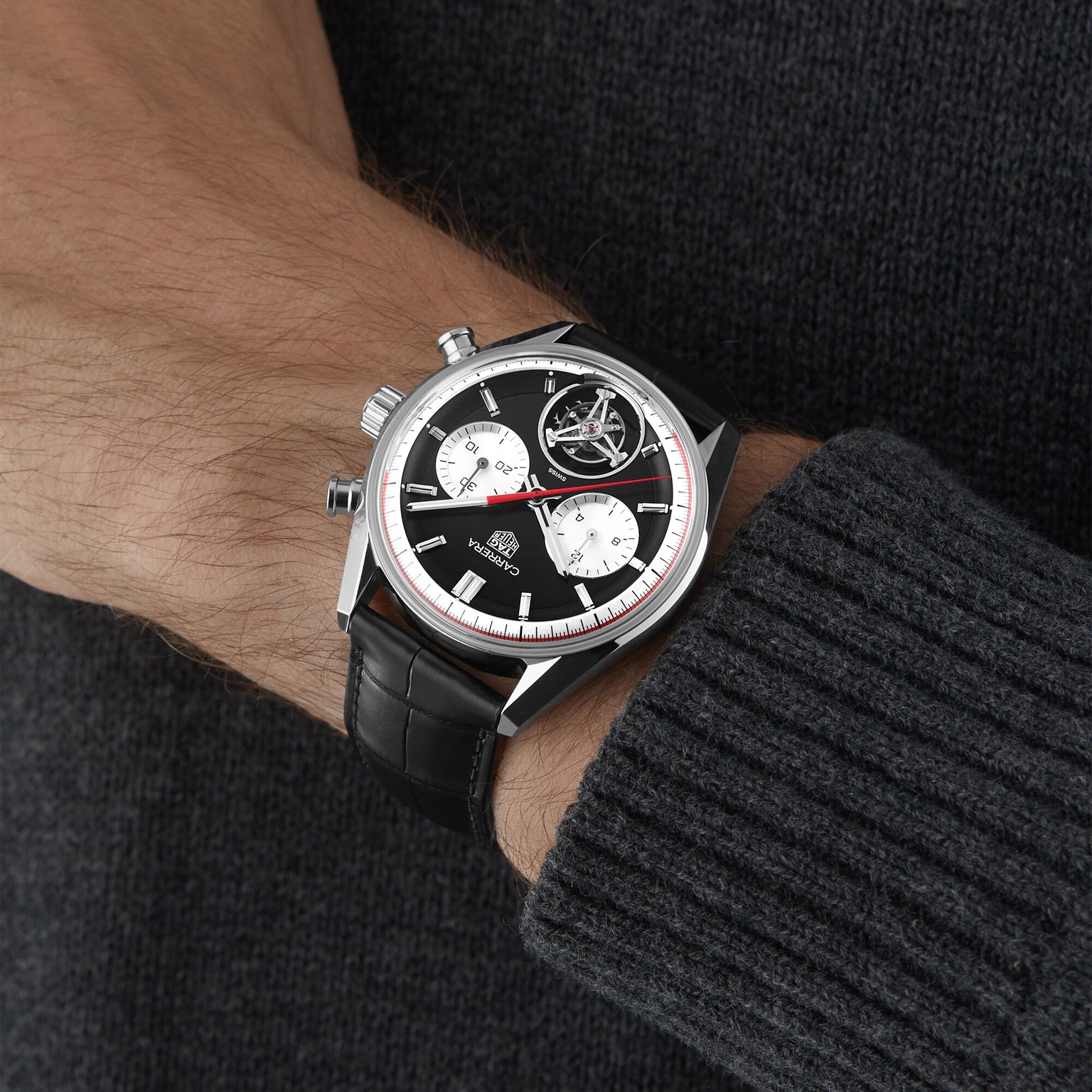 A person wearing a TAG Heuer Carrera Tourbillon Centenary The Watches of Switzerland Group Exclusive chronograph wristwatch with a black leather strap and dark knit sweater.