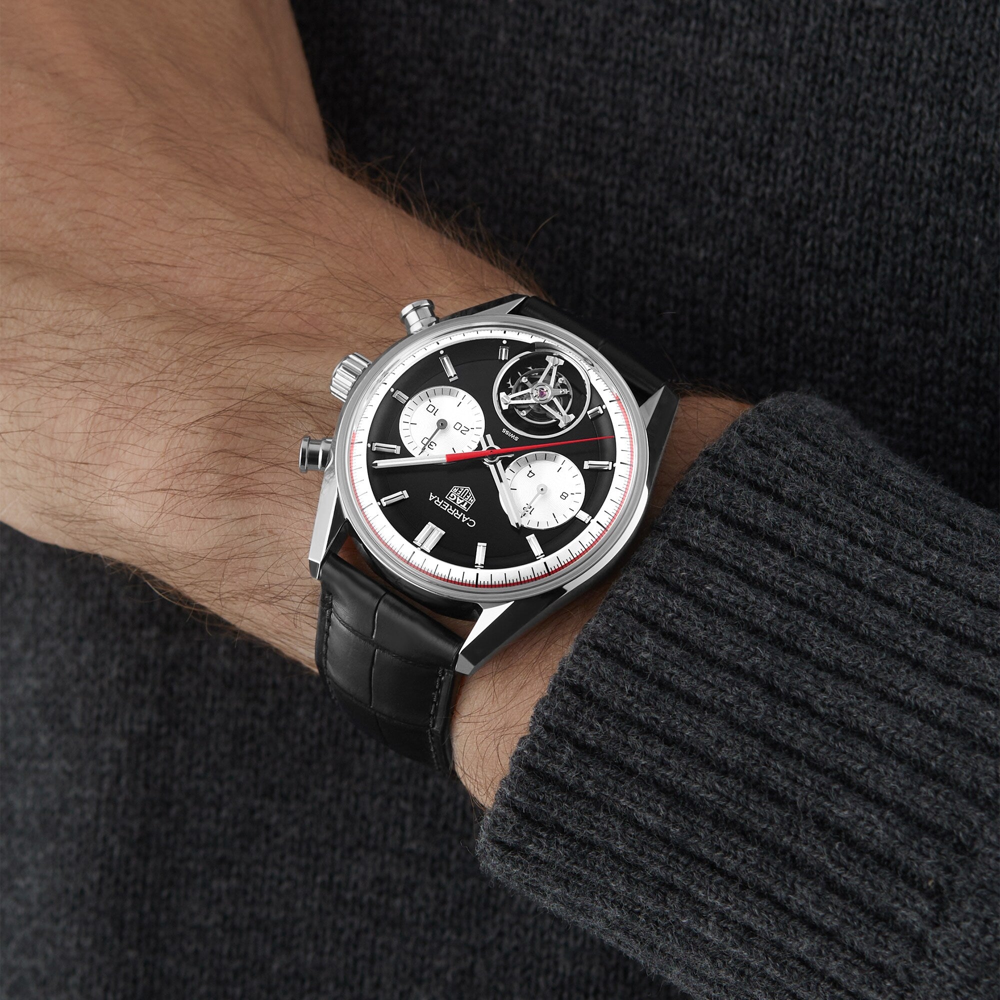 A person wearing a TAG Heuer Carrera Tourbillon Centenary The Watches of Switzerland Group Exclusive chronograph wristwatch with a black leather strap and dark knit sweater.