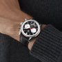 A person wearing a TAG Heuer Carrera Tourbillon Centenary The Watches of Switzerland Group Exclusive chronograph wristwatch with a black leather strap and dark knit sweater.