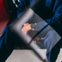 A person in a blue suit sits with hands clasped, wearing the TAG Heuer Carrera Chronosprint x Porsche Rallye Limited Edition, seen through a glass pane.