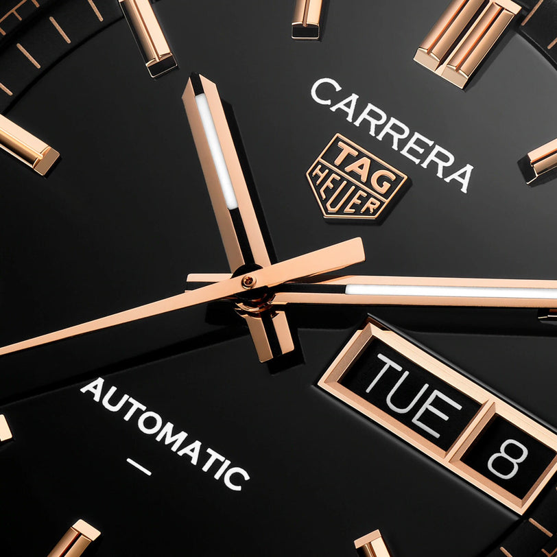 Close-up of the TAG Heuer Carrera Day-Date watch face, featuring gold hands and day-date displays on a black background.