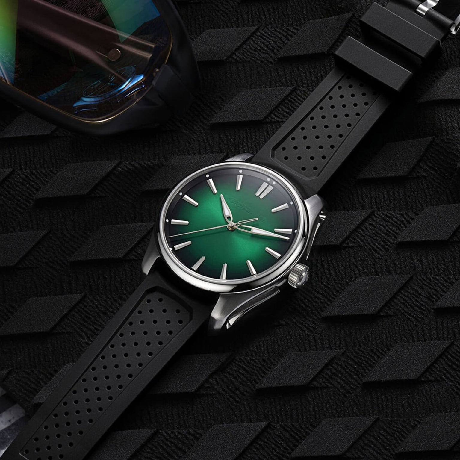 The H. Moser & Cie Pioneer Centre Seconds Cosmic Green wristwatch with a green dial and black strap rests on a textured black surface next to a pair of sunglasses.
