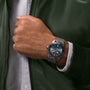 A person in a green jacket and white shirt wears a Breitling Superocean Heritage B20 Automatic 42 with a silver mesh band, blue dial, luminescent hands, and bezel.