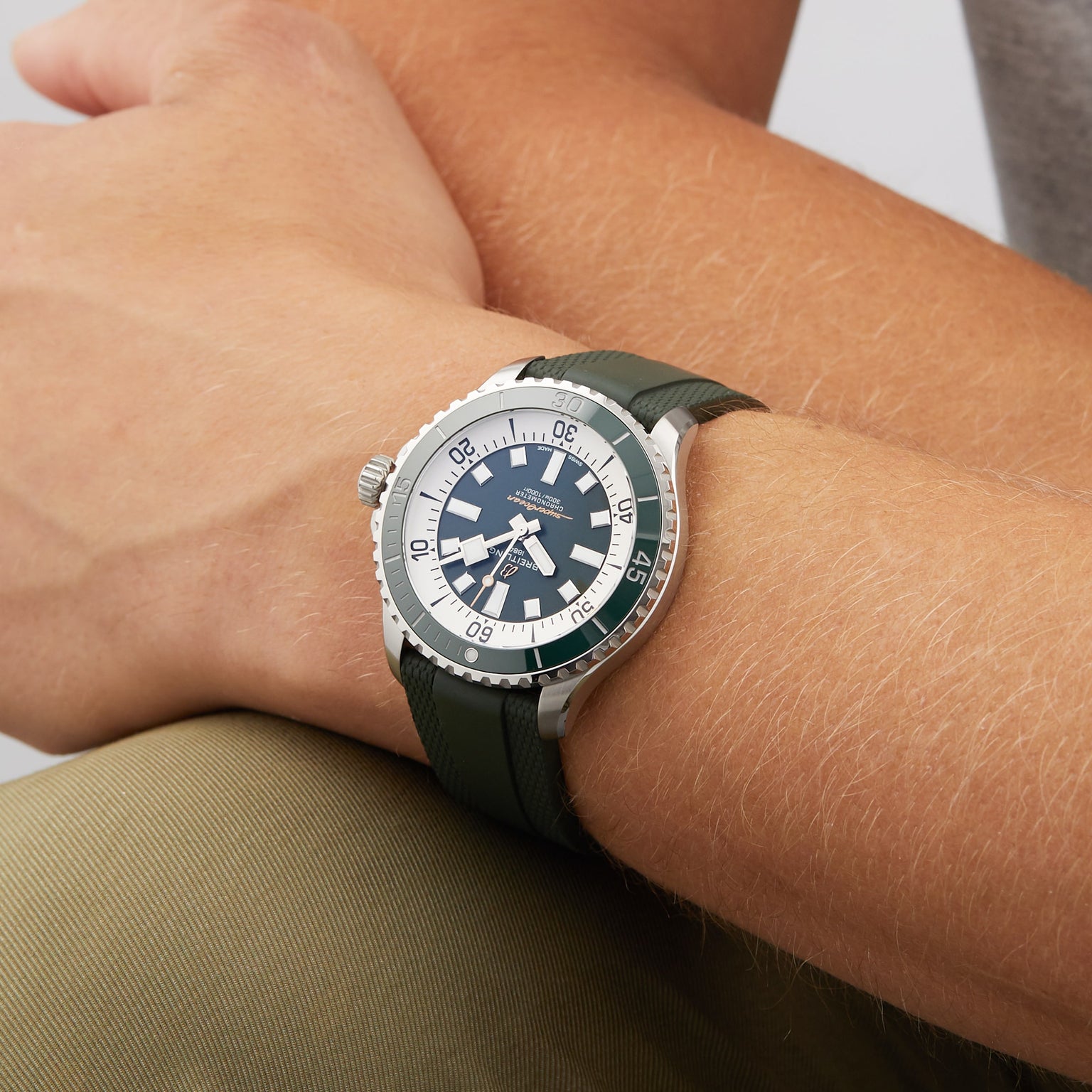 A close-up of a person wearing the Breitling Superocean Automatic 44, a water-resistant silver and black diving watch with a green strap, on their left wrist, while their right hand rests on their left arm.