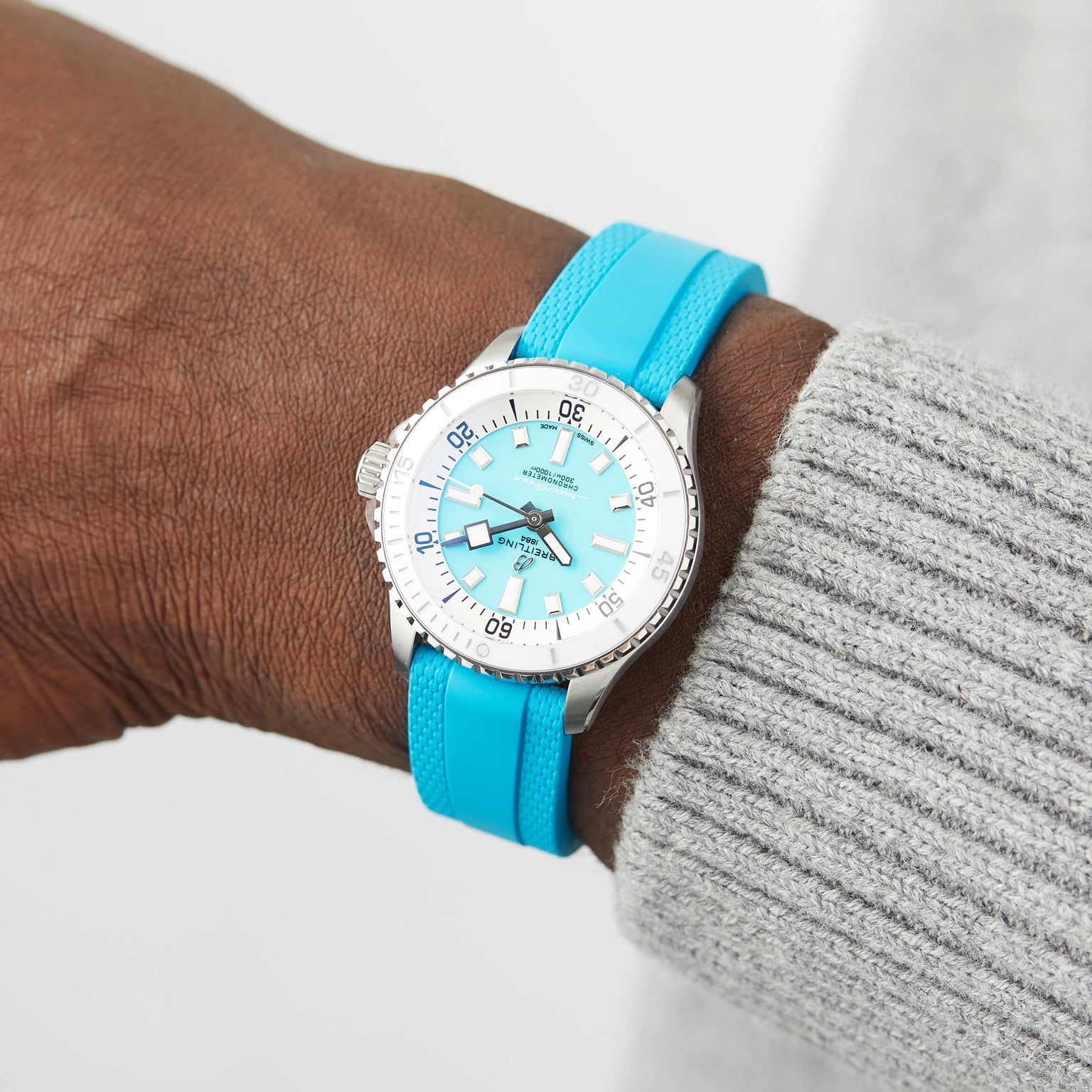 A close-up of a wrist wearing the Breitling Superocean Automatic 36, featuring a silver and turquoise ceramic inlaid bezel and a matching turquoise strap, beside a gray knitted sleeve.
