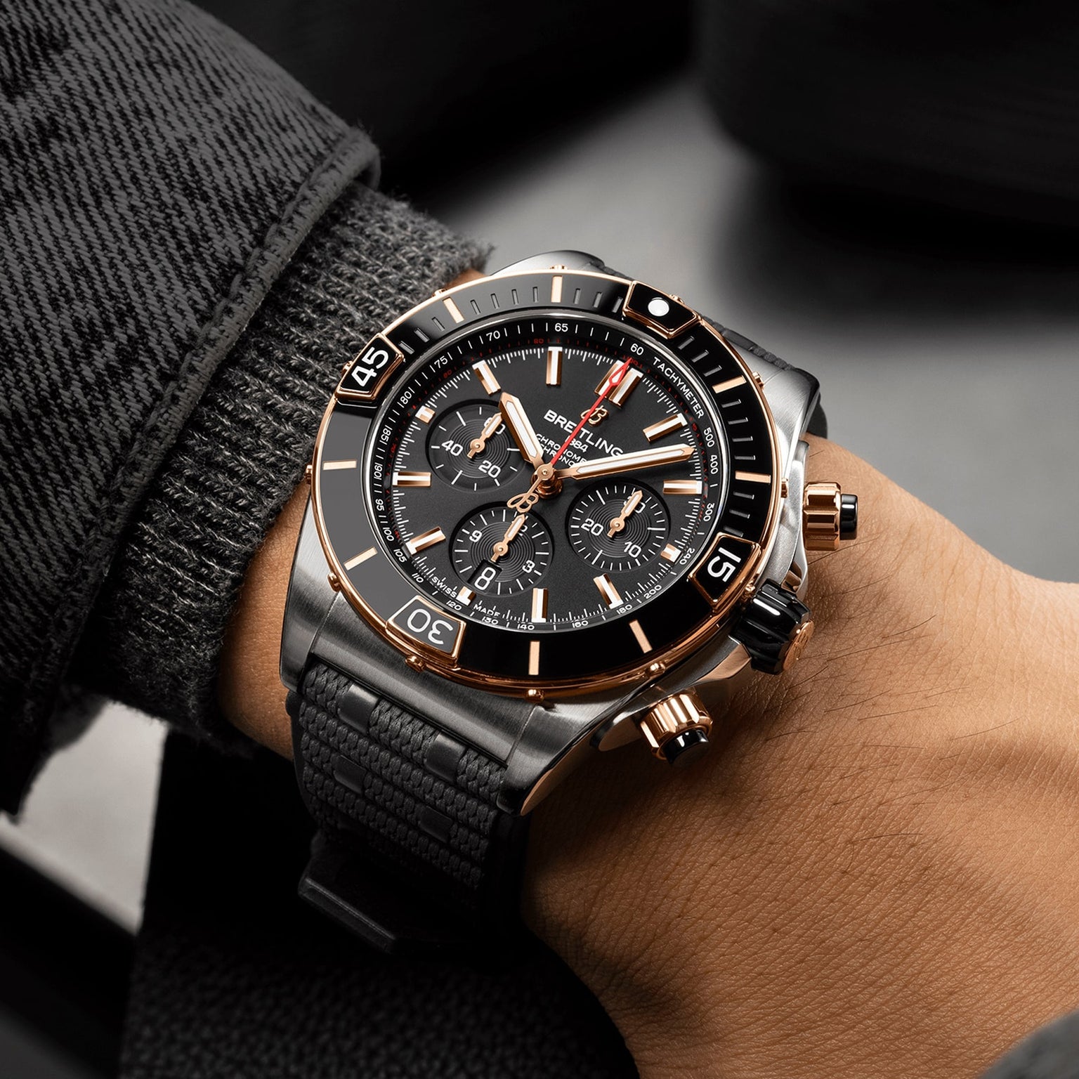 A close-up of a person wearing a Breitling Super Chronomat B01 44 mechanical chronograph with a black strap and textured sleeve visible.