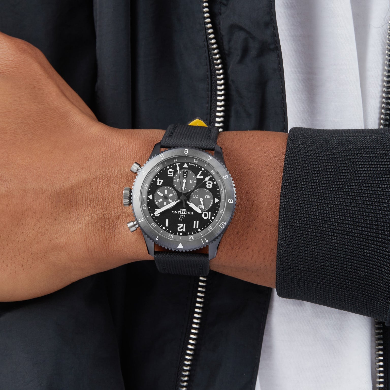 A person in a black jacket and white shirt showcases their left wrist adorned with the Breitling Super AVI B04 Chronograph GMT Mosquito Night, featuring a black strap and multiple dials, inspired by the legacy of the de Havilland Mosquito.