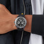 A person in a black jacket and white shirt showcases their left wrist adorned with the Breitling Super AVI B04 Chronograph GMT Mosquito Night, featuring a black strap and multiple dials, inspired by the legacy of the de Havilland Mosquito.