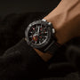 A Breitling Avenger B01 Chronograph 44 Night Mission Dust Devils wristwatch with a black fabric strap and red stitching is worn, showing the time and two subdials on a dark background, powered by the Breitling Manufacture Caliber 01.