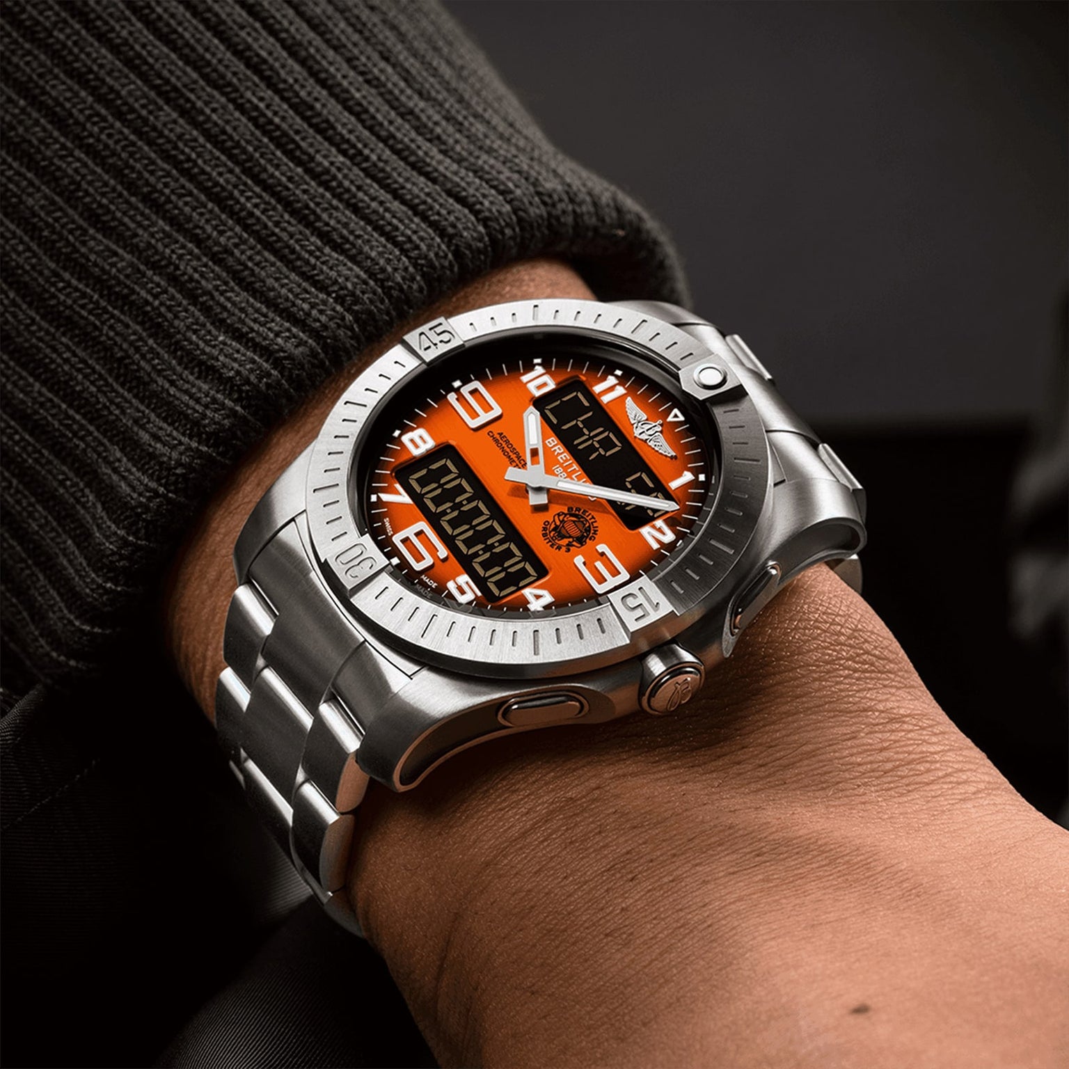 A person wearing a Breitling Aerospace B70 Orbiter wristwatch with a silver case, orange and black dial, digital-analog display, metal bracelet, and pilot’s chronograph features.
