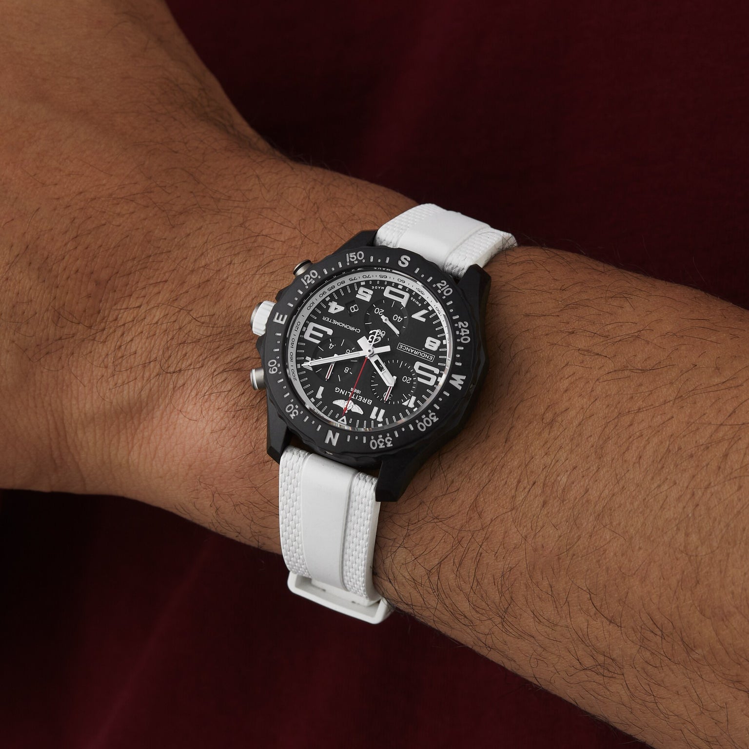 A person wearing a Breitling Endurance Pro 38, a black athleisure watch with white markings and a white band, on their left wrist against a dark red background.