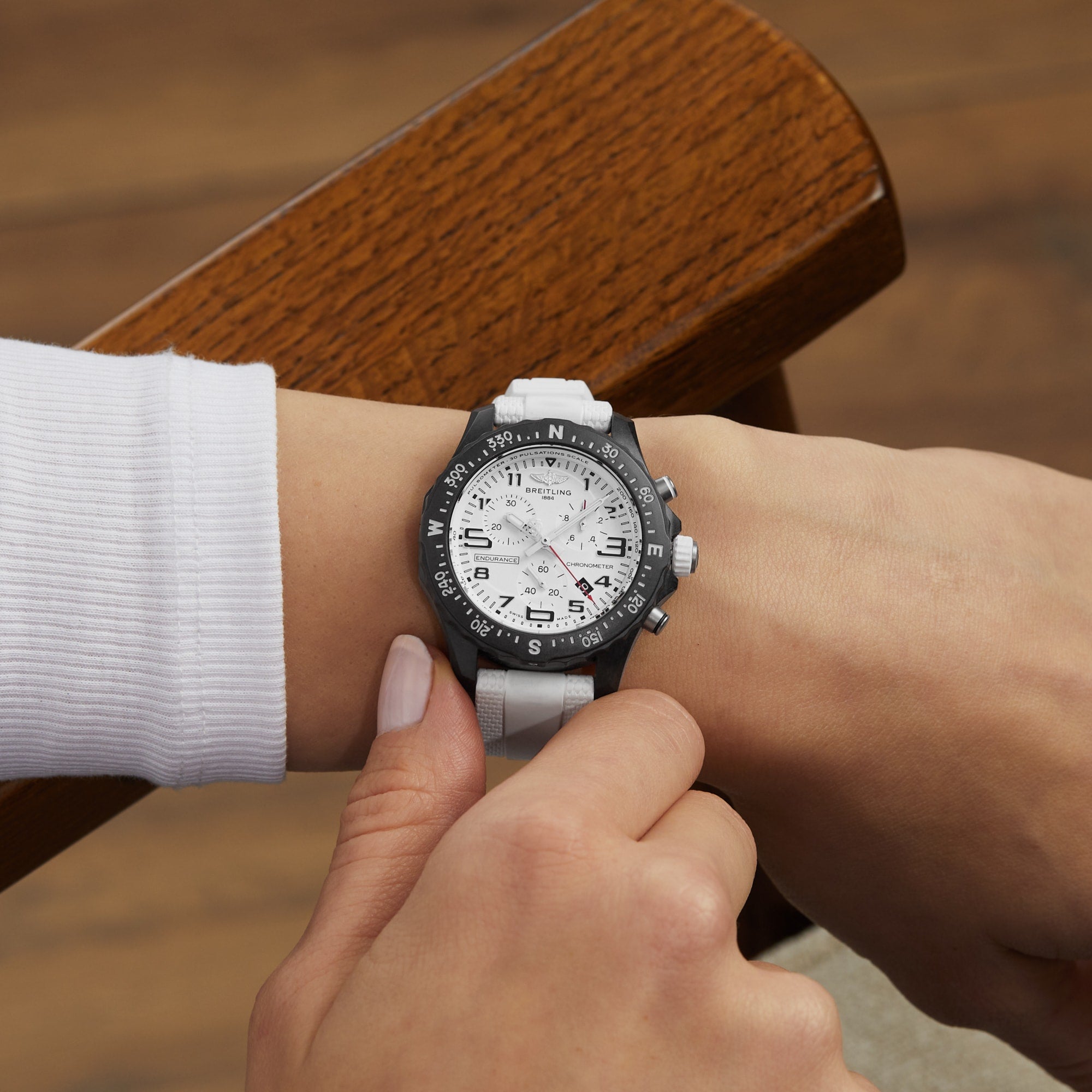 A person wearing the Breitling Endurance Pro 38 The Watches Of Switzerland Group Exclusive, featuring a white strap and black bezel, adjusts it while seated on a wooden chair—showcasing refined style.