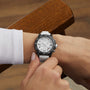 A person wearing the Breitling Endurance Pro 38 The Watches Of Switzerland Group Exclusive, featuring a white strap and black bezel, adjusts it while seated on a wooden chair—showcasing refined style.