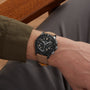 A person wearing the Breitling Avenger B01 Chronograph 42 Night Mission with a tan strap, seated on a wooden chair.