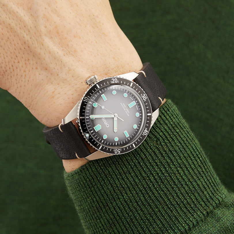 The Oris Divers Sixty‑Five wristwatch, featuring a black strap and glowing hour markers, is worn on the wrist of someone in a green sweater.