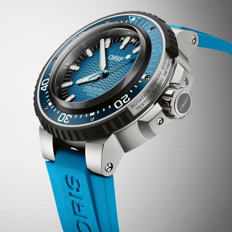 A close-up of the Oris Aquis Pro 4000M dive watch with a blue wave-pattern dial, black bezel, blue strap, Calibre 400 movement, Rotation Safety System, and “Tested to 100 Bar/1000 m” on the case.