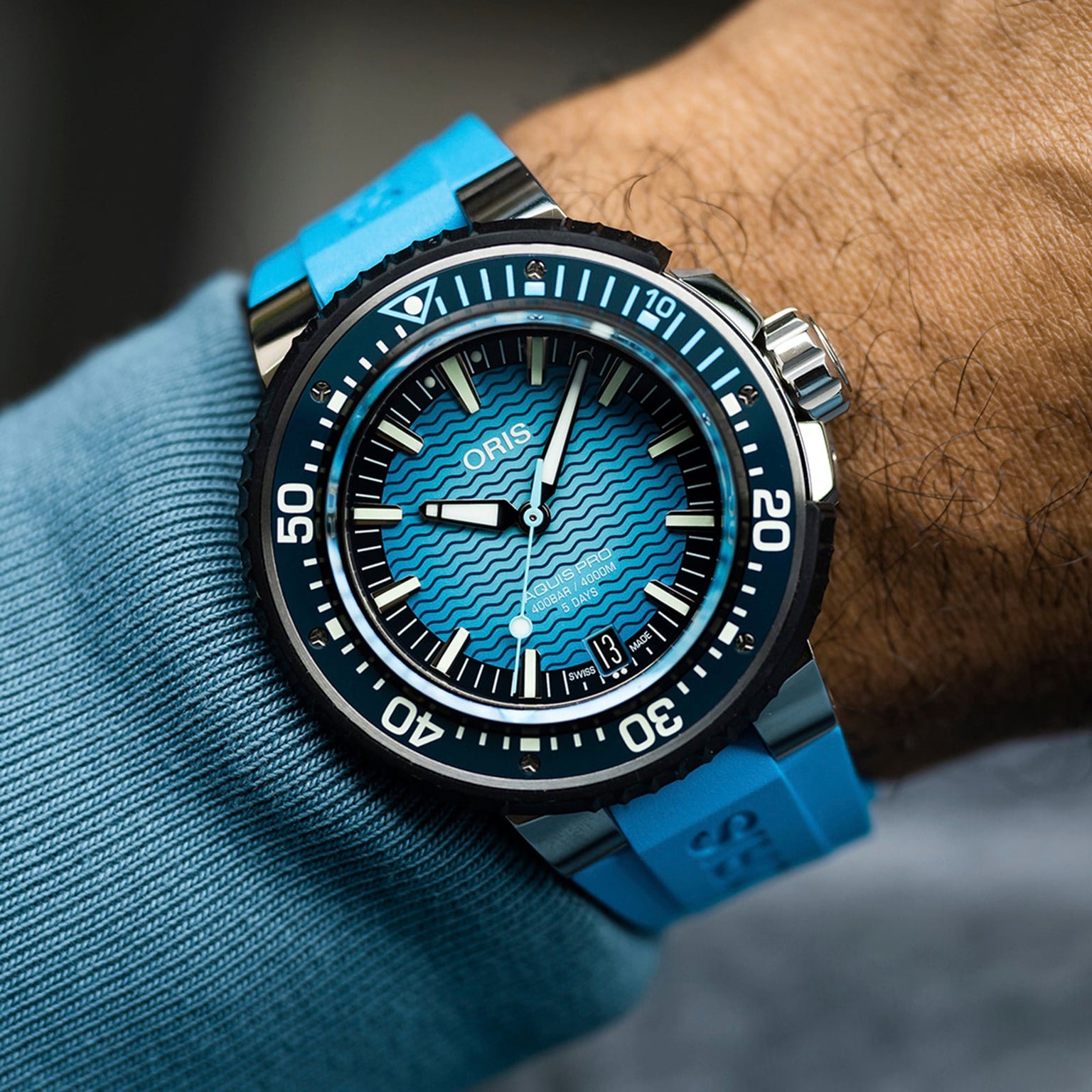 A close-up of a wrist wearing the Oris Aquis Pro 4000M watch, featuring a blue wave-patterned dial, bold white markers, blue rubber strap, and Oris’s trademark Rotation Safety System.