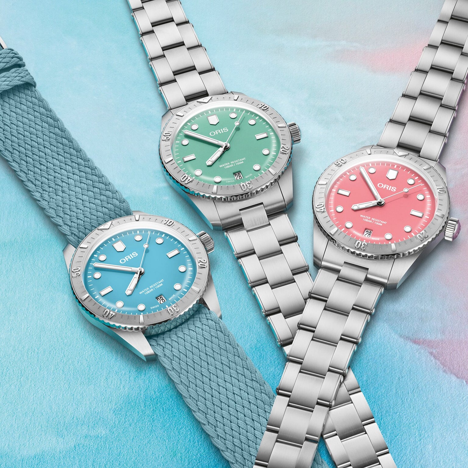 Three Oris Divers Sixty‑Five Cotton Candy watches with blue, green, and pink dials are displayed on a pastel background; one features a fabric strap.