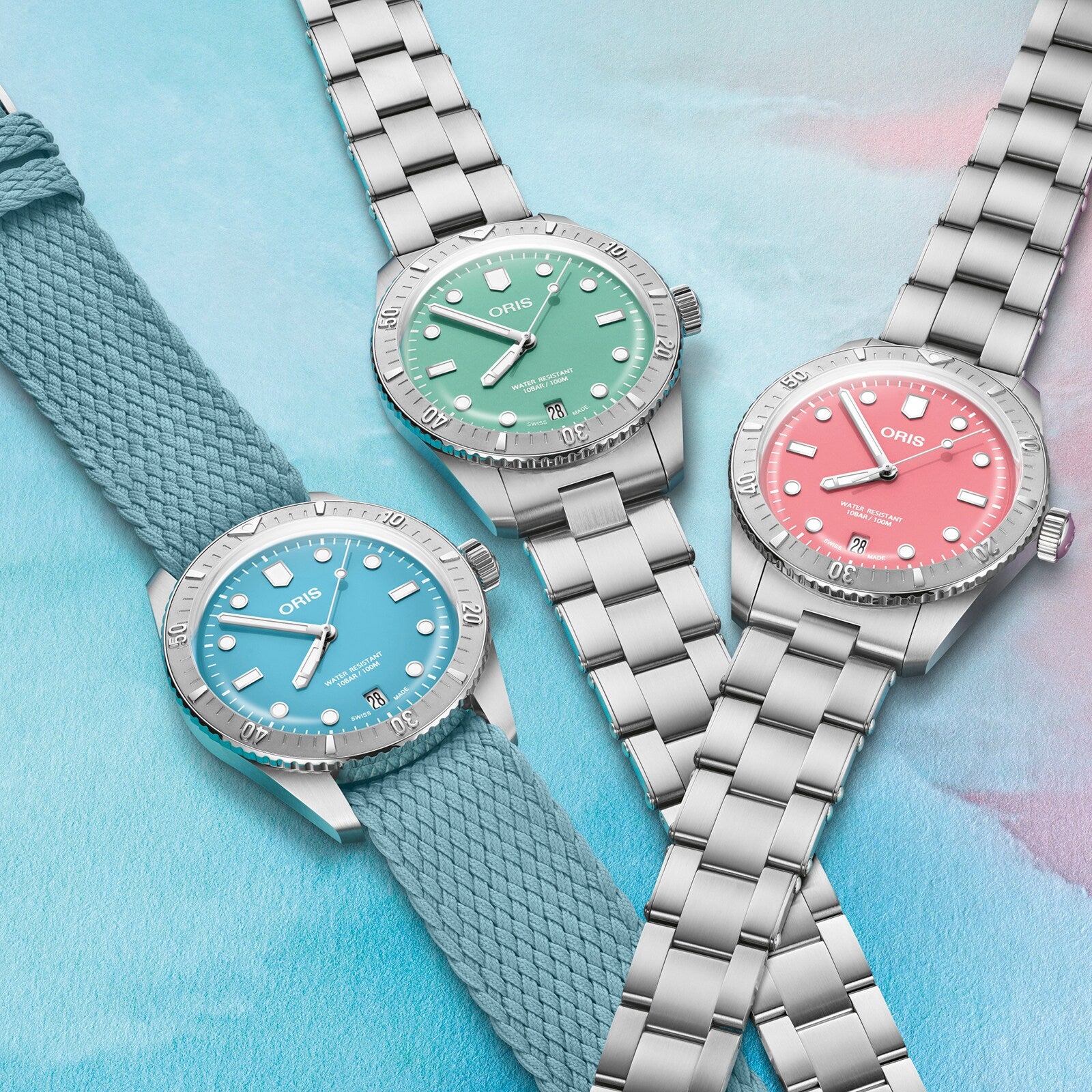 Three Oris Divers Sixty‑Five Cotton Candy watches with blue, green, and pink dials are displayed on a pastel background; one features a fabric strap.