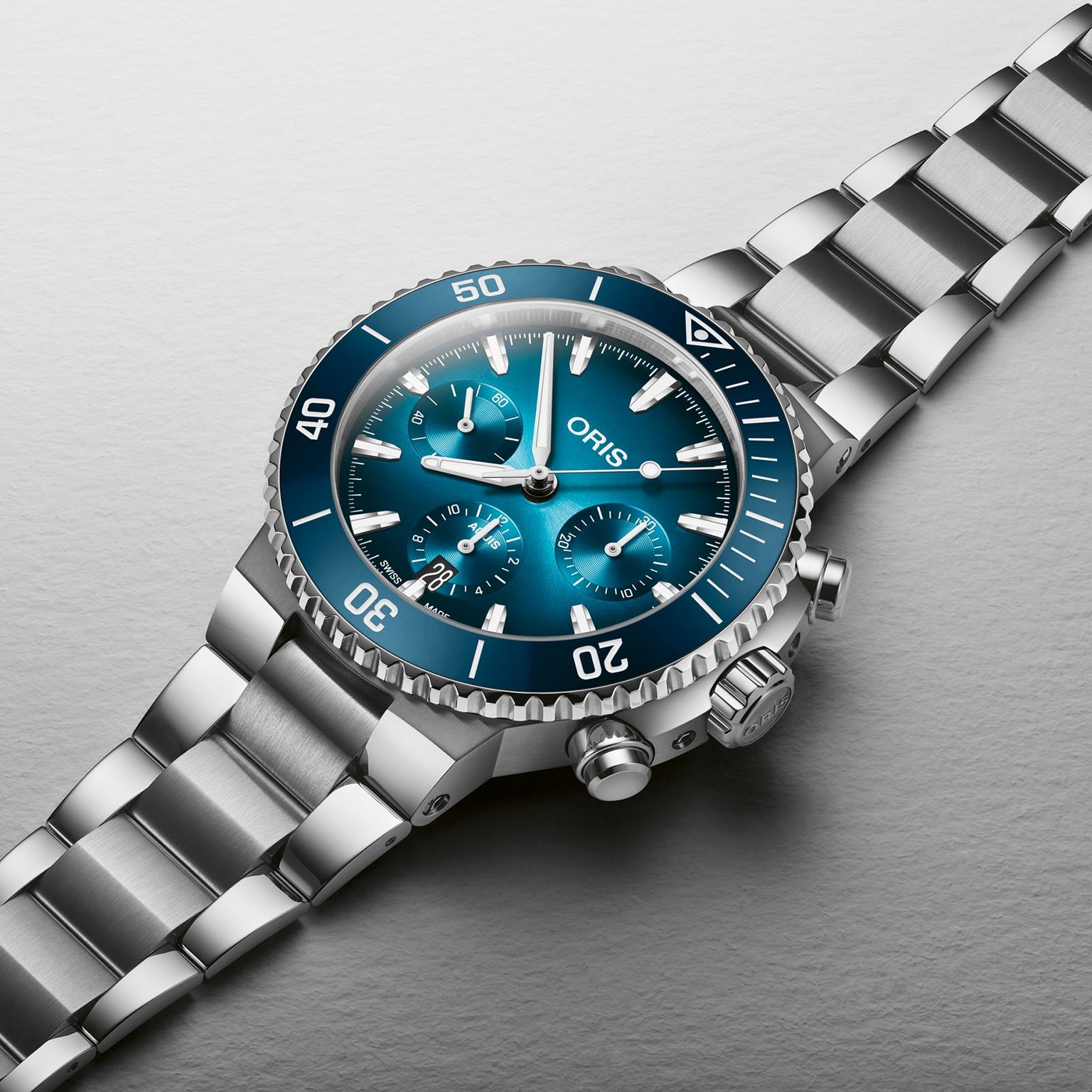 The Oris Aquis Chronograph, a stainless steel wristwatch with a blue dial, ceramic bezel, three subdials, and metallic bracelet, is displayed on a light gray surface to showcase its exceptional water resistance.
