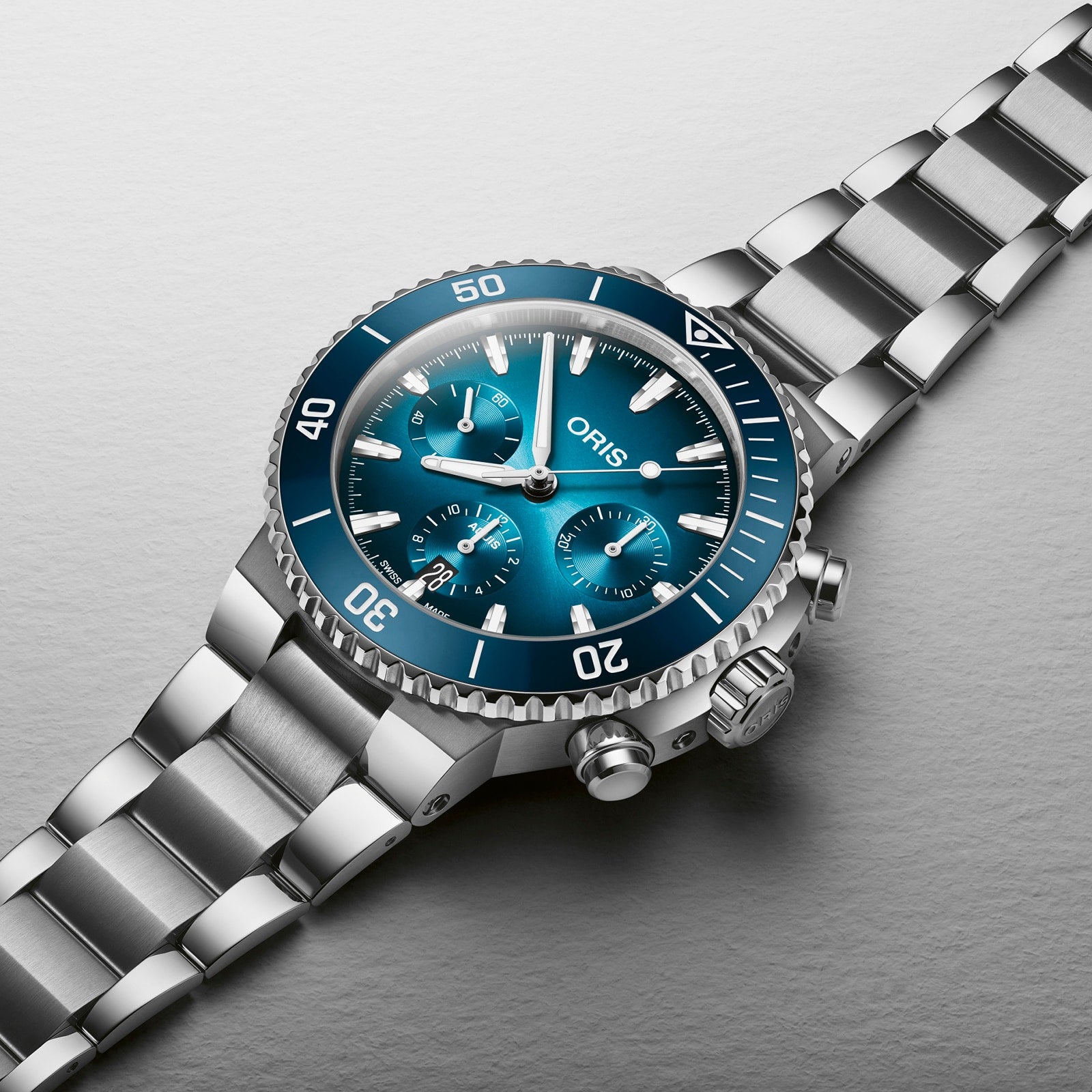 The Oris Aquis Chronograph, a stainless steel wristwatch with a blue dial, ceramic bezel, three subdials, and metallic bracelet, is displayed on a light gray surface to showcase its exceptional water resistance.