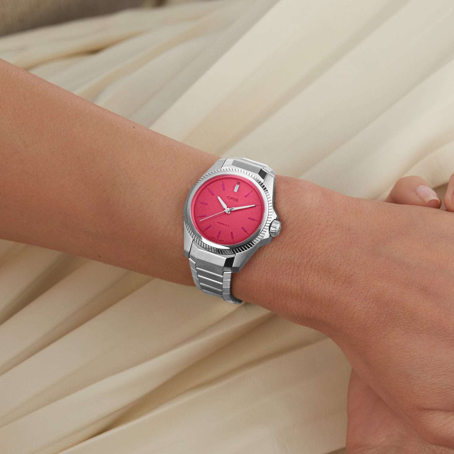 A person’s wrist wears an Oris ProPilot X Miss Piggy Edition watch with a hot pink dial and silver case, set against cream-colored fabric.