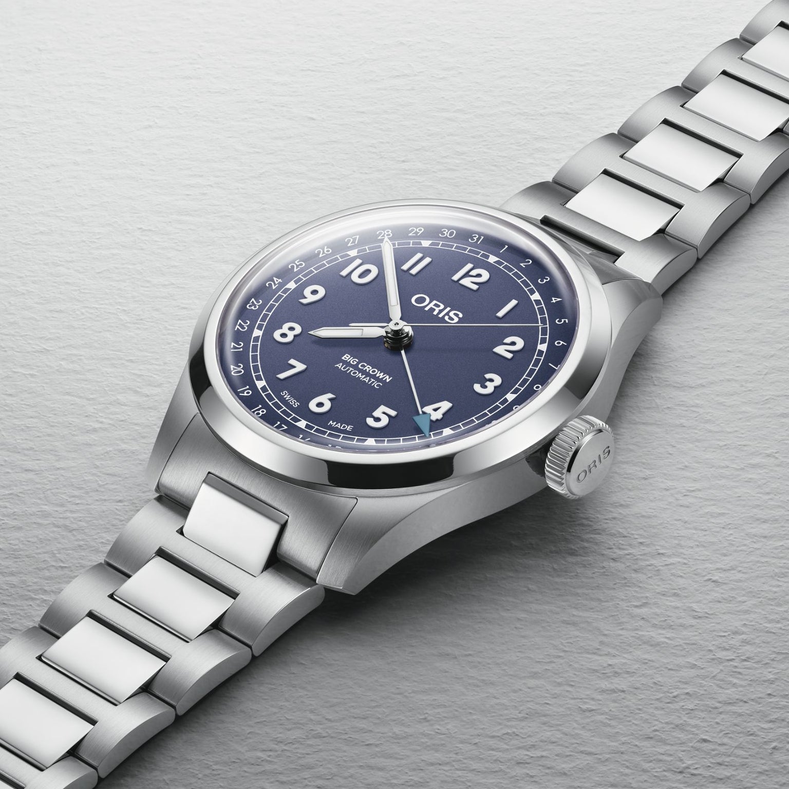 An Oris Big Crown Pointer Date wristwatch with a blue dial, white Arabic numerals, metal bracelet, and Swiss Made automatic movement is displayed on a light textured surface.