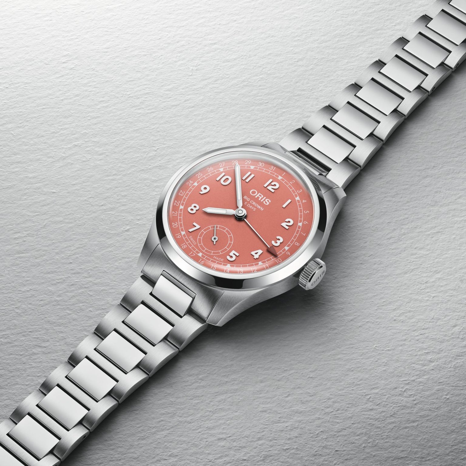 The Oris Big Crown Pointer Date Calibre 403, featuring a stainless steel case, pink dial with white numerals, and metal bracelet, is displayed on a textured light gray background.
