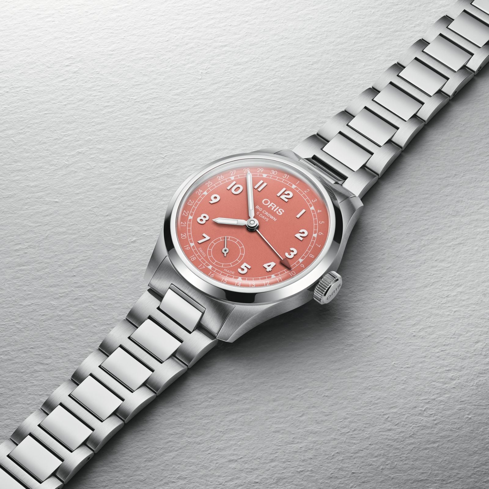 The Oris Big Crown Pointer Date Calibre 403, featuring a stainless steel case, pink dial with white numerals, and metal bracelet, is displayed on a textured light gray background.