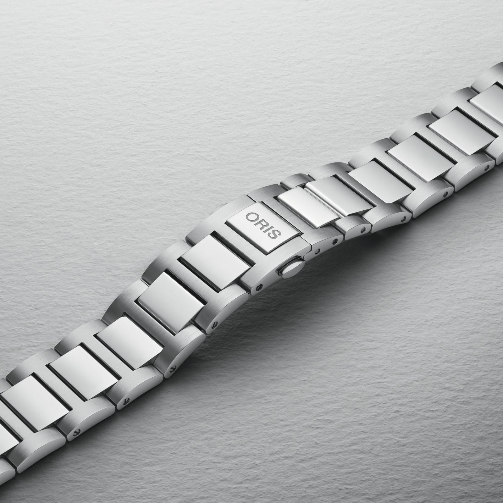 The Oris Big Crown Diamonds watch features a stainless steel bracelet with a brushed finish and an engraved clasp, showcased against a textured light gray background to emphasize its diamond hour markers.