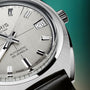 Silver-toned automatic watch with square date window, black hour markers, and textured dial against teal background.