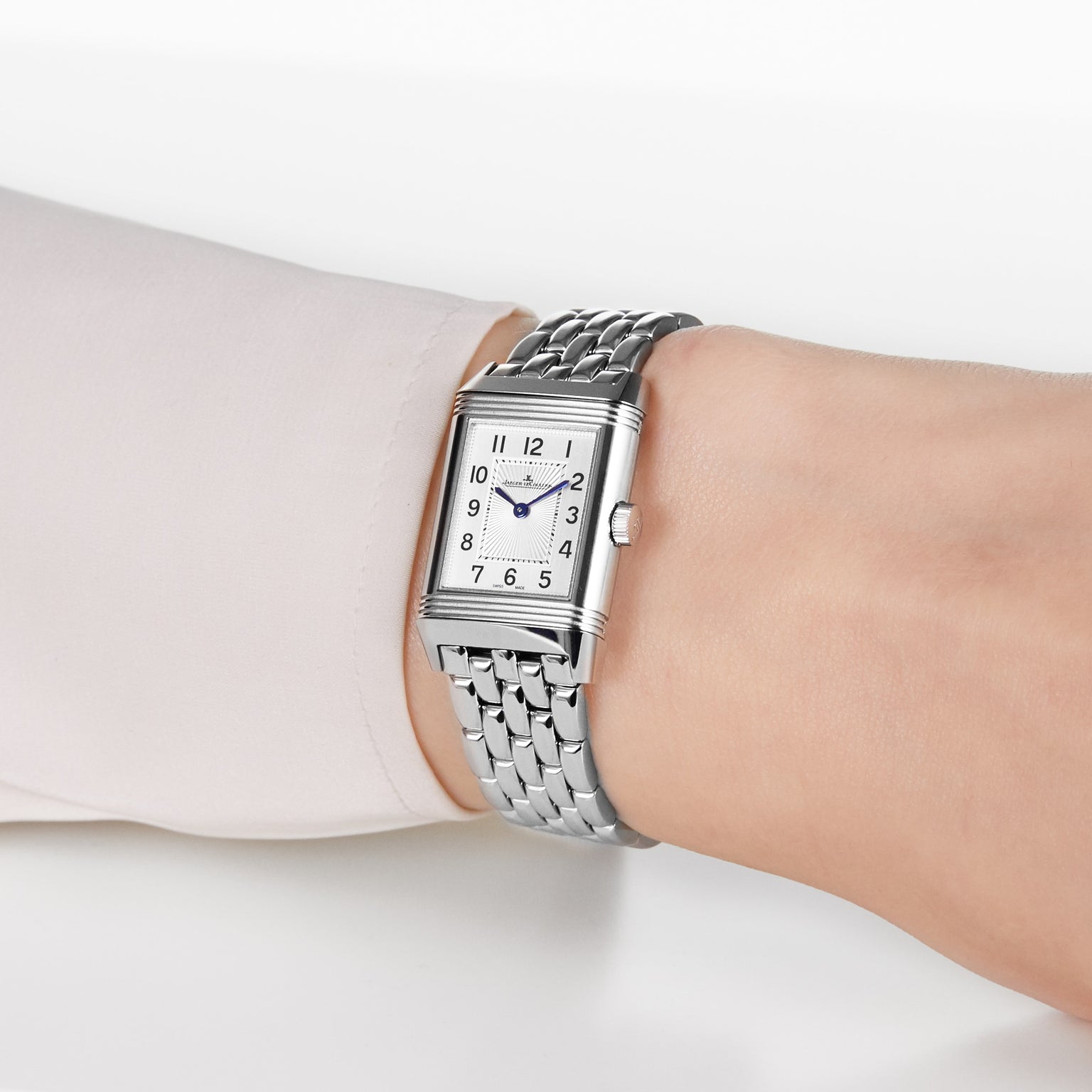 A close-up of a wrist wearing the Jaeger-LeCoultre Reverso Classic, featuring a silver rectangular case, metal link band, off-white dial with black numerals and blue hands. The diamond bezel complements a light-colored long-sleeve shirt.