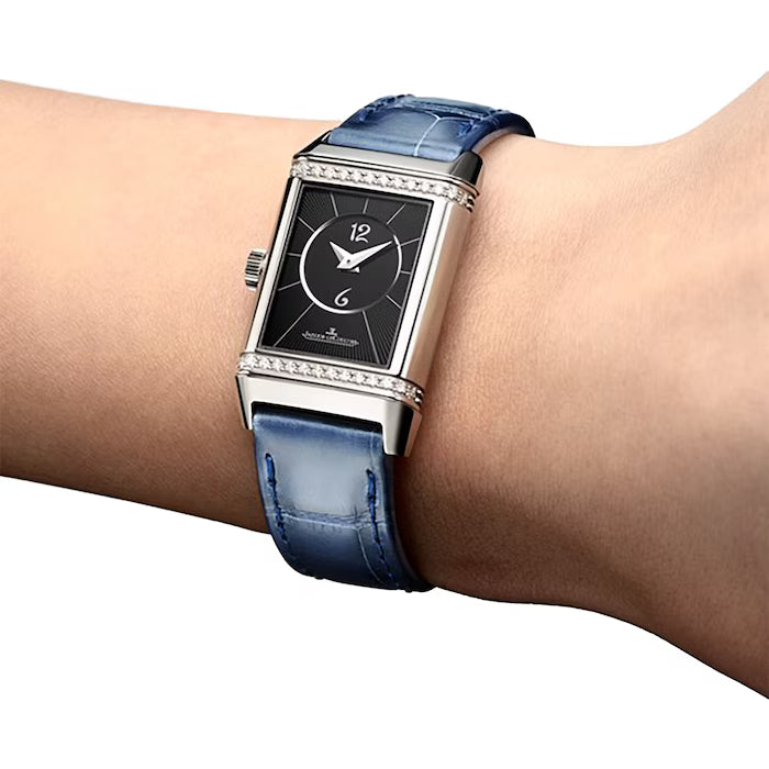 A Jaeger-LeCoultre Reverso Small Duetto wristwatch with a rectangular black face, silver details, and a blue leather strap is worn on a person’s wrist, highlighting its minimalist design.