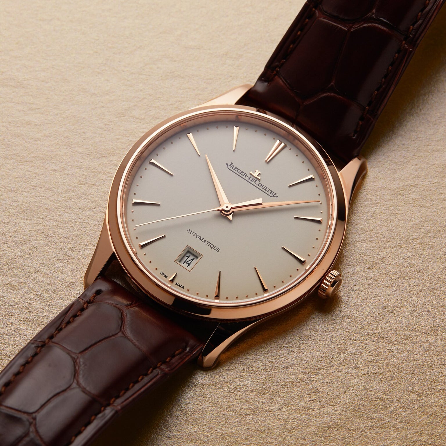 The Jaeger-LeCoultre Master Ultra Thin Date has a brown leather strap, pink gold case, white dial with simple hour markers, date window at 6 o’clock, and runs on Caliber 899. The watch is set against a textured beige background.