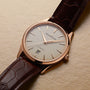 The Jaeger-LeCoultre Master Ultra Thin Date has a brown leather strap, pink gold case, white dial with simple hour markers, date window at 6 o’clock, and runs on Caliber 899. The watch is set against a textured beige background.