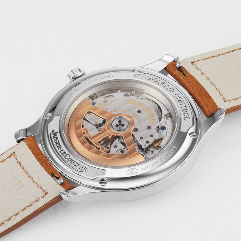 A close-up of the back of the Jaeger-LeCoultre Master Control Date watch reveals its intricate automatic movement through a transparent stainless steel case back, showcasing visible gears, a gold rotor, and a tan leather strap.