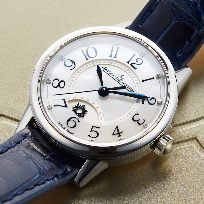 The Jaeger-LeCoultre Rendez-Vous Night & Day is a luxury women’s watch featuring a blue leather strap, white dial with black Arabic numerals, diamond hour markers, blue hands, and a sun-and-moon subdial near 10 o’clock.