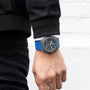 A person in black jacket and pants is shown from the waist down, wearing a Zenith Defy 21 Ultrablue wristwatch with a blue strap and dark detailed face on their left wrist.