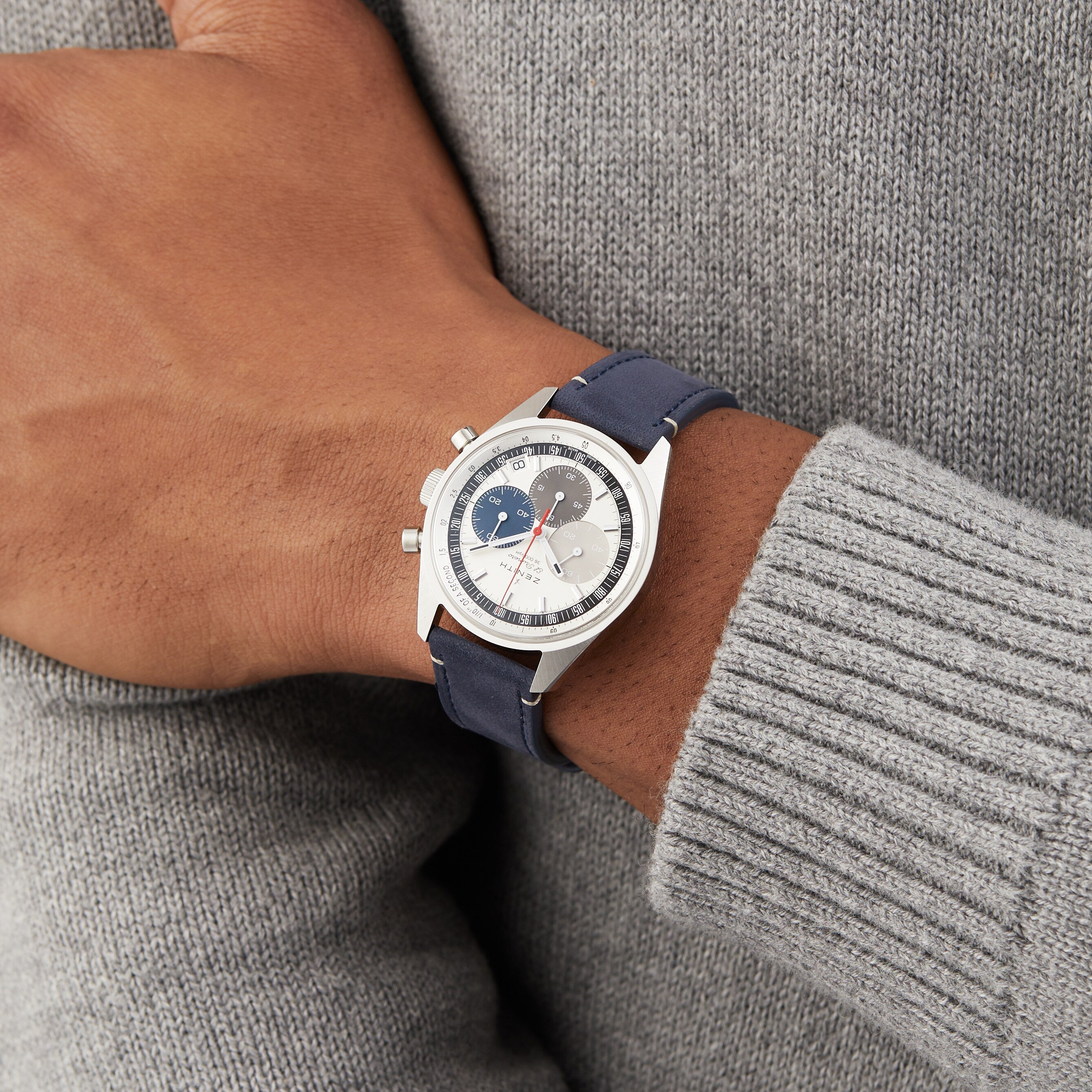 A person in a gray sweater showcases the Zenith Chronomaster EP Original, featuring a silver case, blue strap, and a round white tricolour dial with blue and gray subdials.