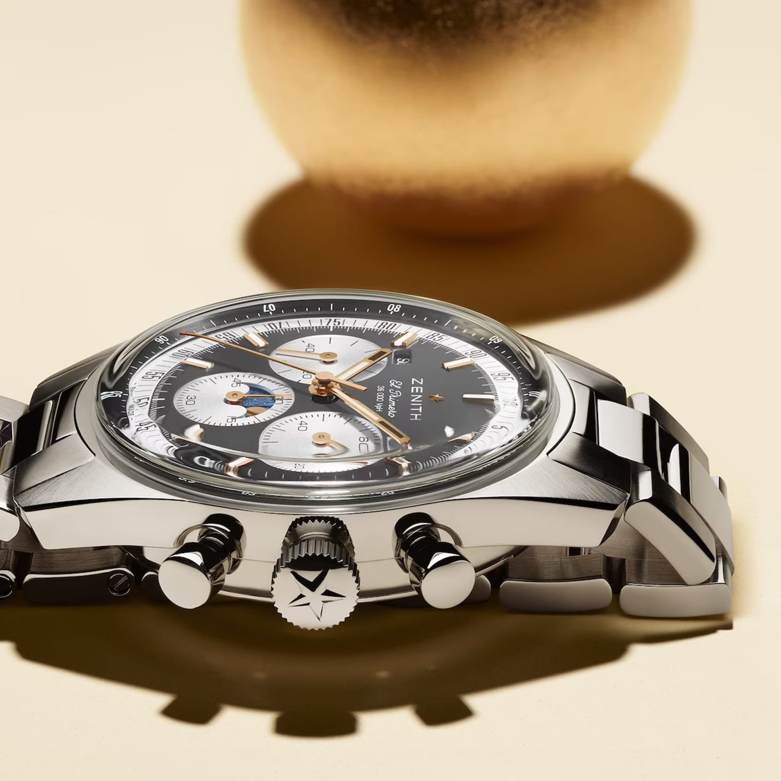 A Zenith Chronomaster Original stainless steel chronograph wristwatch with a black bezel, silver and white subdials, and a metal bracelet is shown on a light surface with a blurred gold object in the background.