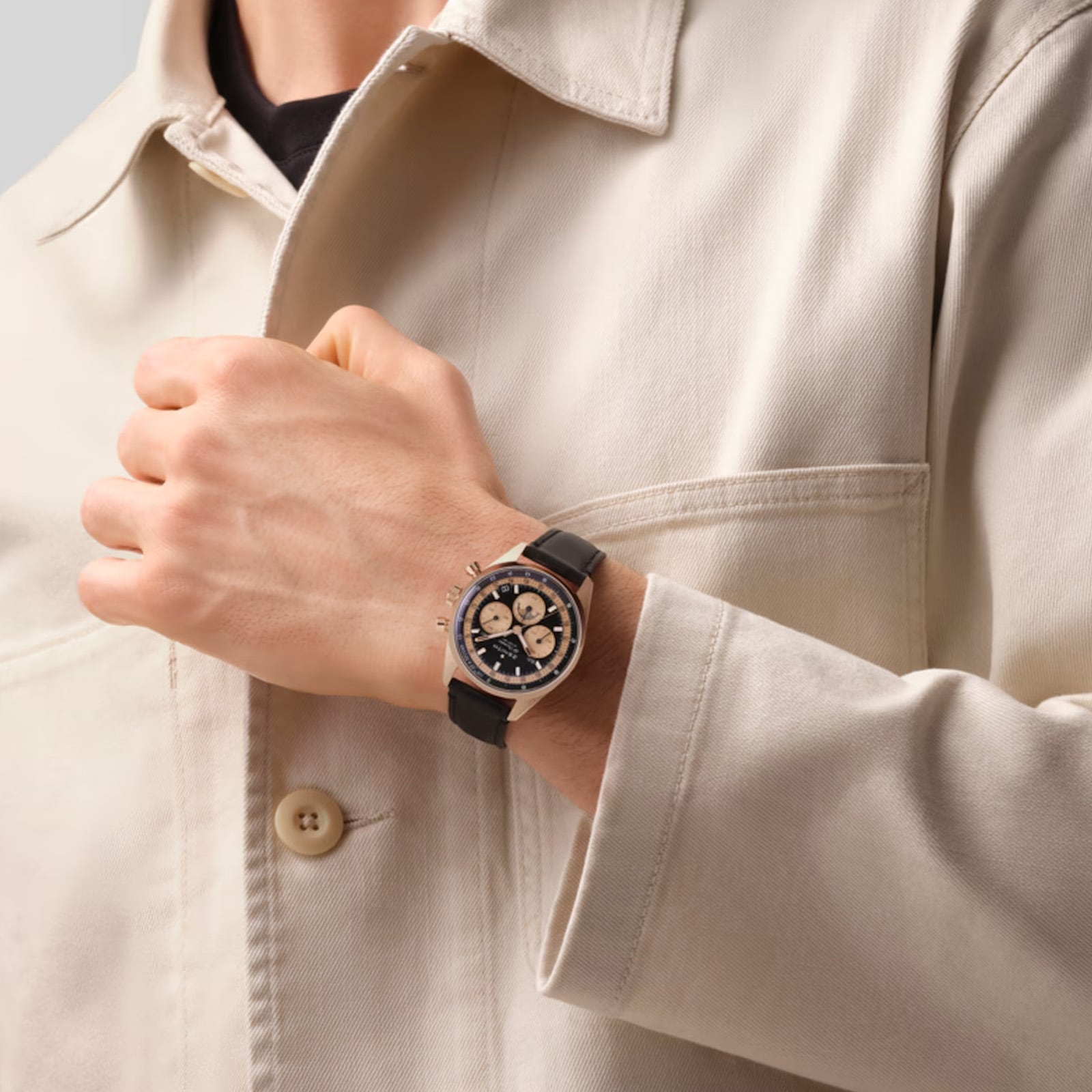 A person in a beige jacket is wearing a Zenith Chronomaster Original, a black and silver watch with a black strap, on their left wrist.