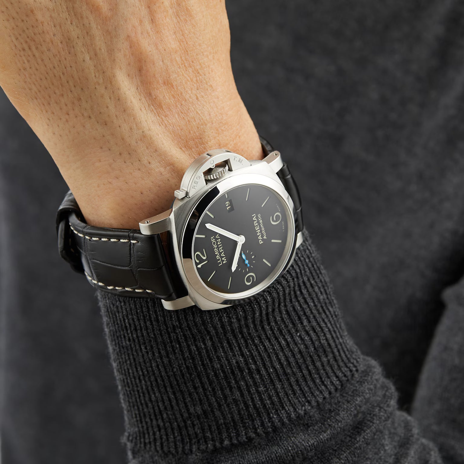 A person in a black sweater wears the Panerai Luminor Marina, an Italian-designed luxury watch with a black leather strap, large round face, silver bezel, and bold white numerals and hands.