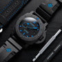 A Panerai Submersible Carbotech wristwatch with blue accents rests on a black surface beside a black strap, Panerai tool, and logo case—highlighting its robust carbon fibre build for divers.
