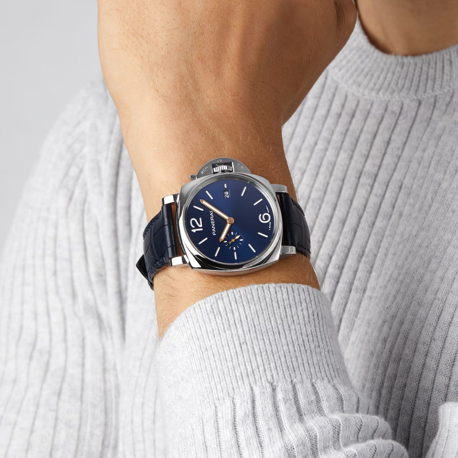 A person in a light gray ribbed sweater showcases a Panerai Luminor Due with a blue dial, silver case, and dark blue leather strap on their left wrist.