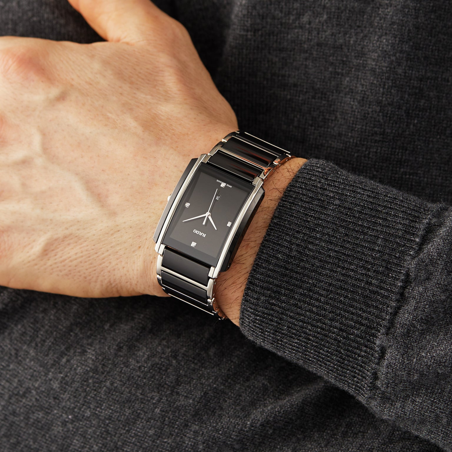A person wearing a stylish Rado Integral Diamonds watch with a rectangular black and silver face and metal band, featuring quartz movement, rests their hand on a dark gray sweater.