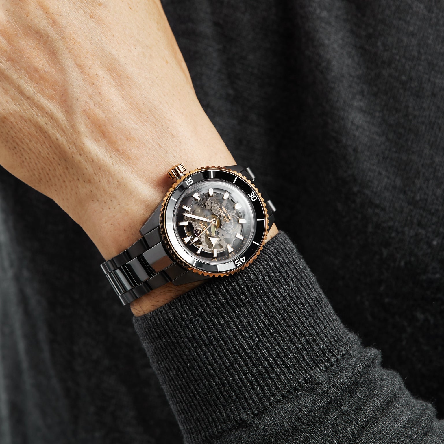 A close-up of a person wearing a Rado Captain Cook High-Tech Ceramic watch with visible mechanical movement and Nivachron™ hairspring on their left wrist, dressed in a dark sweater.